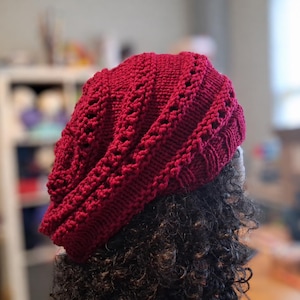Puede incluir: Un gorro de punto de color rojo oscuro con un patrón texturizado de trenzas verticales y puntos calados. El gorro presenta un borde ancho acanalado y una silueta holgada, confeccionado con hilo de construcción tejida visible.