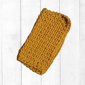 May include: Mustard yellow crocheted item, rectangular in shape. The item has a textured, ribbed appearance, with a slightly scalloped edge. The background is a white, vertical-planked surface.