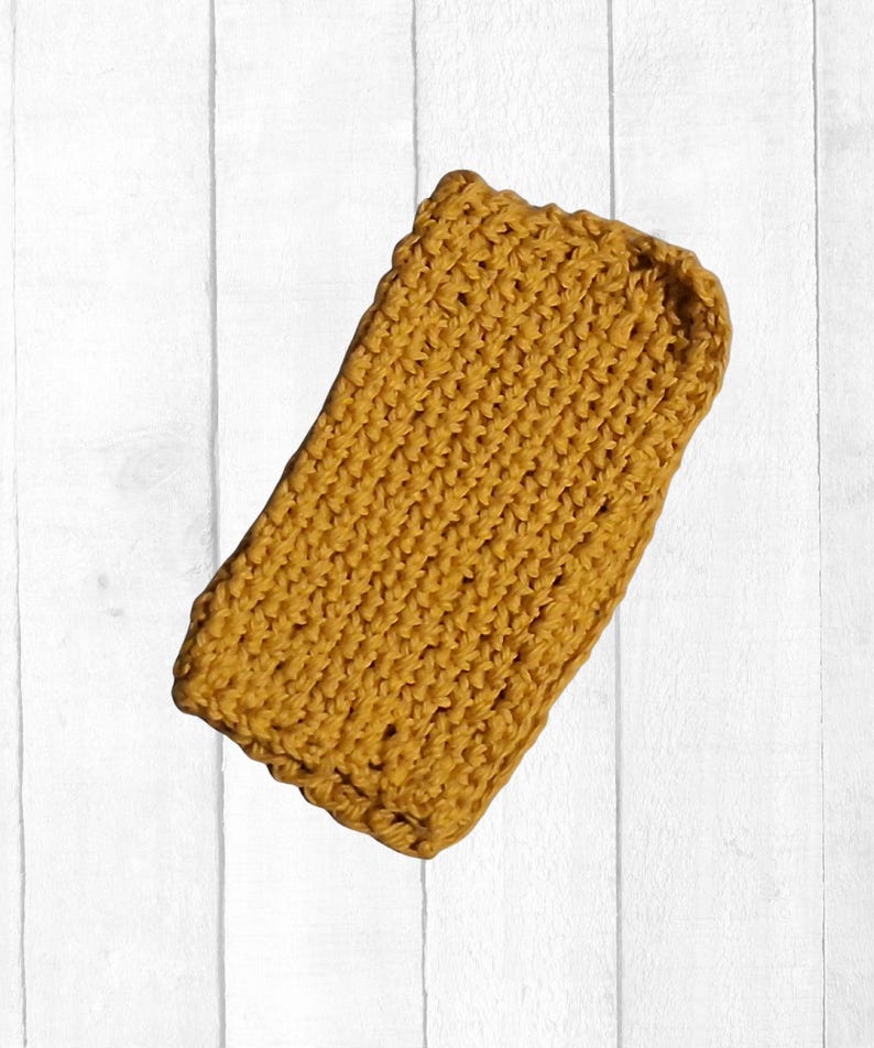 May include: Mustard yellow crocheted item, rectangular in shape. The item has a textured, ribbed appearance, with a slightly scalloped edge. The background is a white, vertical-planked surface.