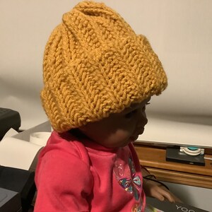 May include: A close-up of a child wearing a mustard yellow knitted beanie hat. The hat has a textured knit pattern and is pulled down over the child's forehead.