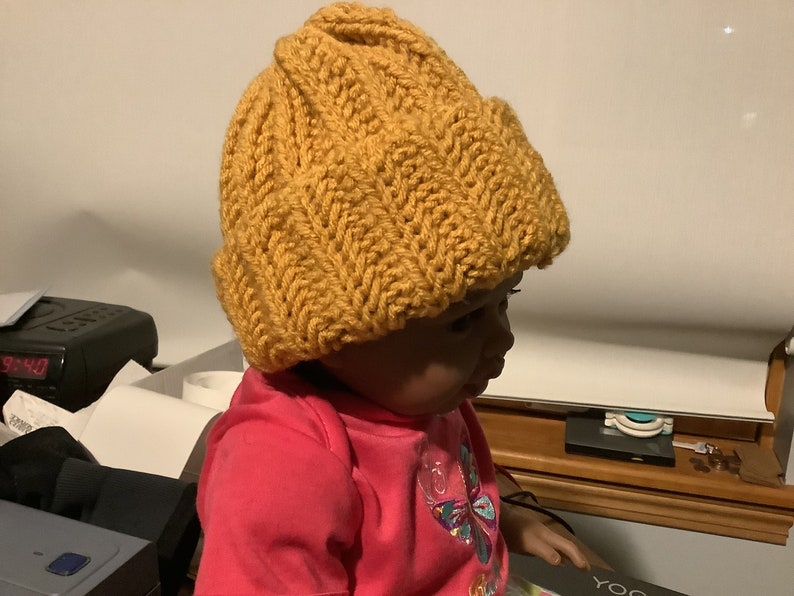May include: A close-up of a child wearing a mustard yellow knitted beanie hat. The hat has a textured knit pattern and is pulled down over the child's forehead.