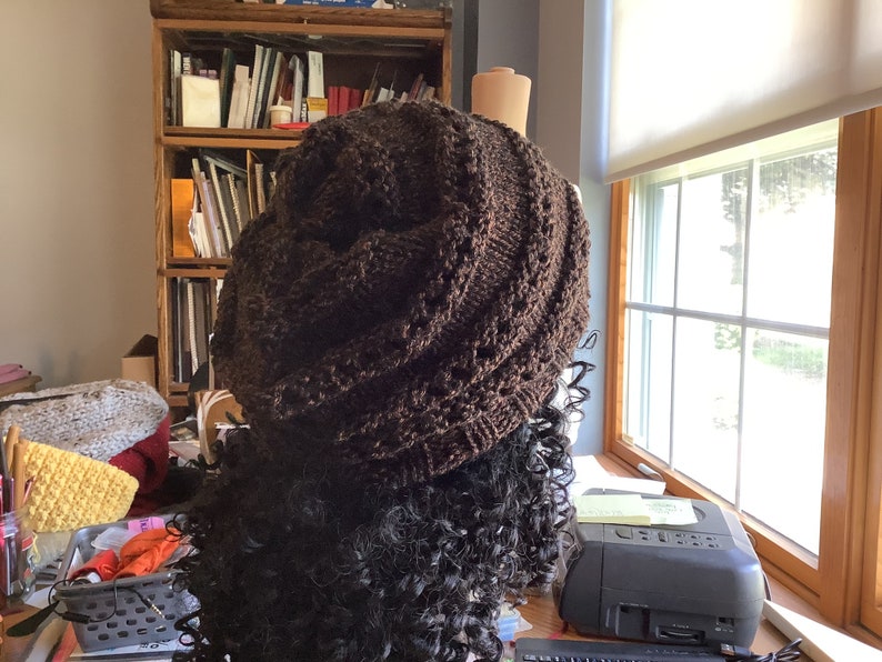 May include: A brown knitted beanie hat with a textured pattern. The hat is on a mannequin head with long, curly, dark brown hair.