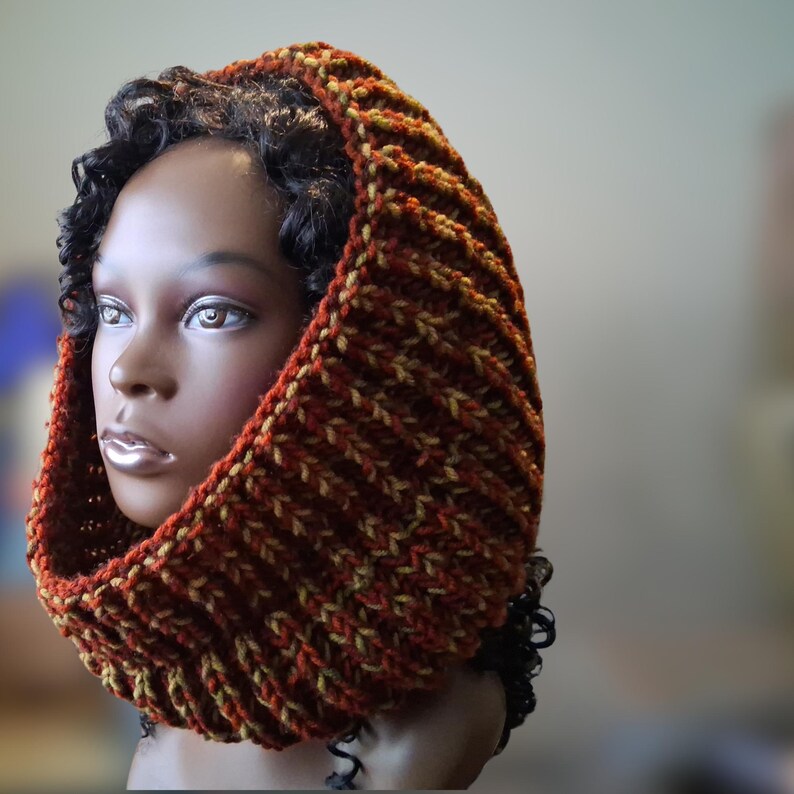 May include: A chunky knit cowl hood featuring a ribbed texture in a variegated yarn blend of deep rust orange, brown, and olive green tones. The thick, hand-knitted accessory frames the head and neck with a warm, dense, and flexible construction.