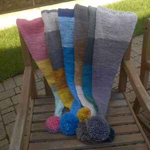 Puede incluir: Cinco bufandas de punto de colores con pompones en un banco de madera. Las bufandas son en tonos de rosa, gris, azul, marr&oacute;n y amarillo.