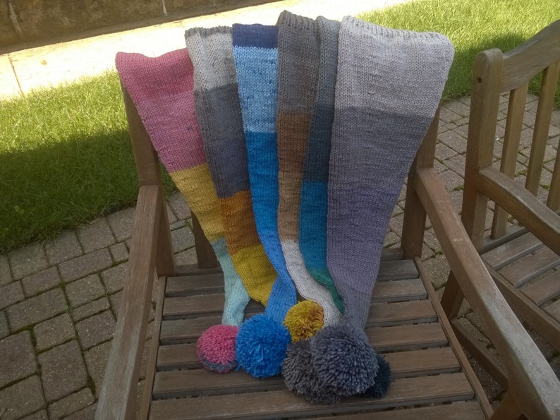 Puede incluir: Cinco bufandas de punto de colores con pompones en un banco de madera. Las bufandas son en tonos de rosa, gris, azul, marr&oacute;n y amarillo.