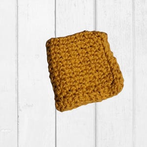 May include: Mustard yellow crocheted square, likely a washcloth or dishcloth, with a textured surface. The square is made of yarn and has a handmade appearance. The background is a white wooden surface.