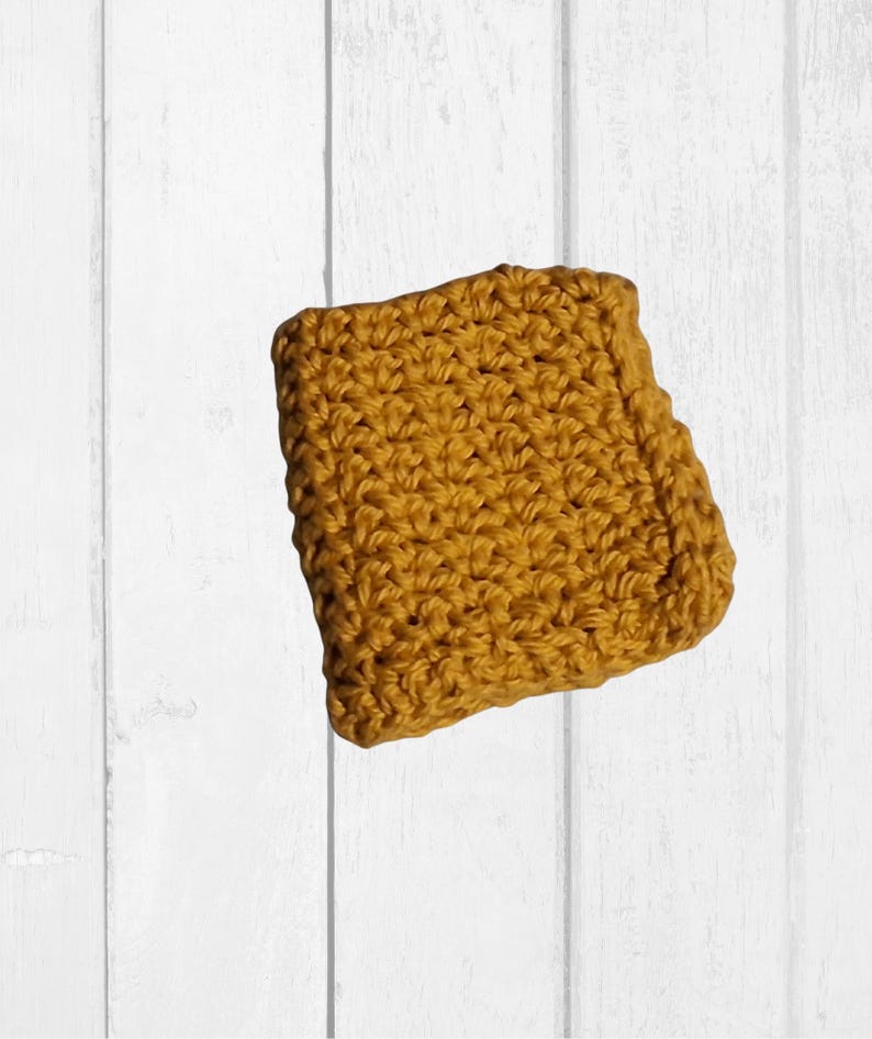 May include: Mustard yellow crocheted square, likely a washcloth or dishcloth, with a textured surface. The square is made of yarn and has a handmade appearance. The background is a white wooden surface.