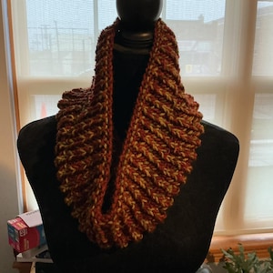 May include: A chunky knit infinity scarf featuring a ribbed texture with a variegated color pattern of deep rust, burnt orange, and muted olive green yarn. The scarf is displayed on a black mannequin, showcasing its thick, circular construction.
