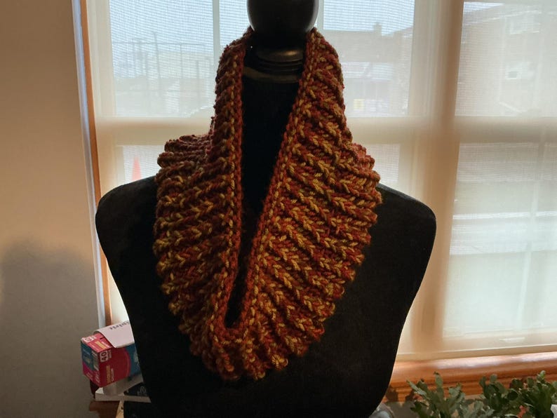 May include: A chunky knit infinity scarf featuring a ribbed texture with a variegated color pattern of deep rust, burnt orange, and muted olive green yarn. The scarf is displayed on a black mannequin, showcasing its thick, circular construction.