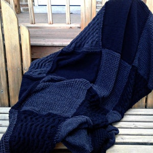 May include: A black and gray knitted throw blanket with a patchwork design. The blanket is folded and draped over a wooden bench.