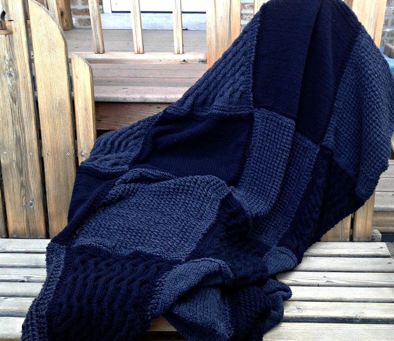 May include: A black and gray knitted throw blanket with a patchwork design. The blanket is folded and draped over a wooden bench.