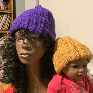 May include: Two knitted hats, one purple and one mustard yellow. The hats are on the heads of two mannequins.