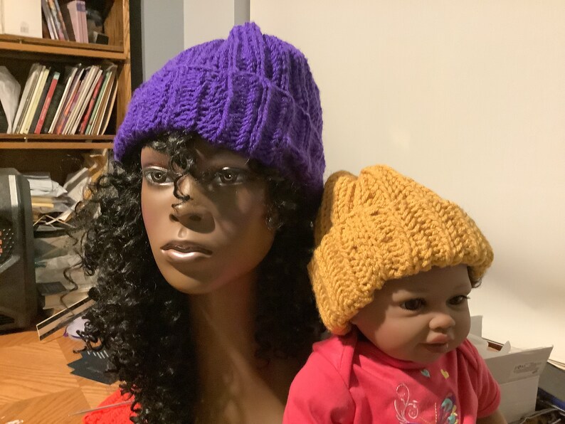 May include: Two knitted hats, one purple and one mustard yellow. The hats are on the heads of two mannequins.