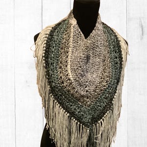 May include: A handmade, fringed shawl in a triangular shape. The shawl features a gradient of colors, transitioning from light beige to shades of gray and green. The fringe is a light beige color.