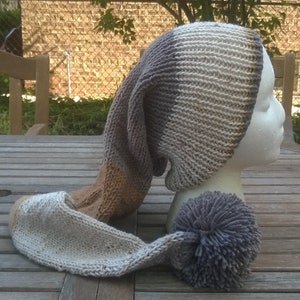 Puede incluir: Un gorro largo de punto con un patr&oacute;n de rayas marrones, grises y blancas. El gorro tiene un gran pomp&oacute;n en el extremo de la cola larga.