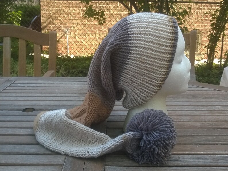 Puede incluir: Un gorro largo de punto con un patr&oacute;n de rayas marrones, grises y blancas. El gorro tiene un gran pomp&oacute;n en el extremo de la cola larga.