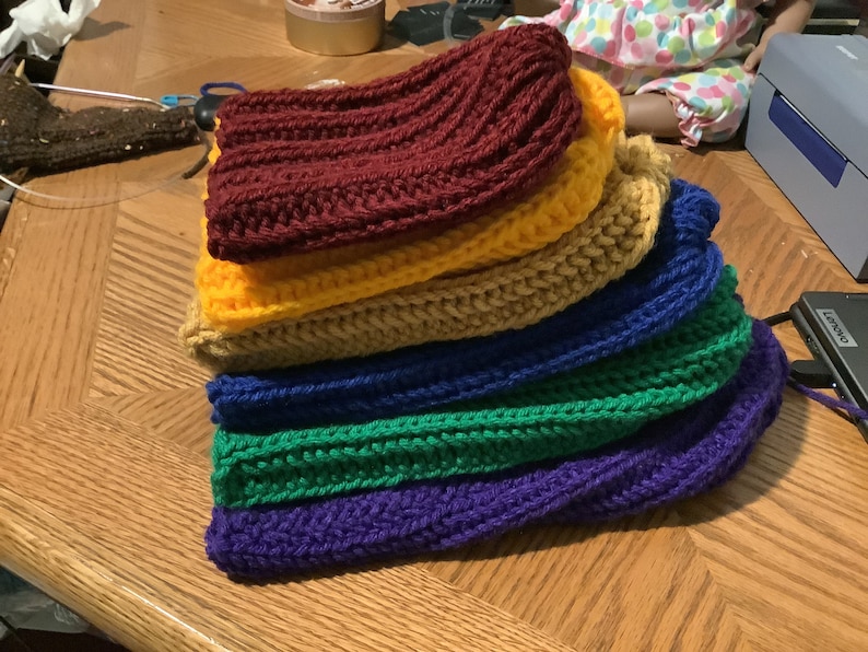 May include: A stack of six crocheted hats in a rainbow color pattern. The hats are made of different colored yarn, starting with a maroon hat at the top and ending with a purple hat at the bottom. The hats are stacked on a wooden surface.