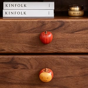 以下が含まれることがあります： 赤とタン色のリンゴ型の木製引き出しノブ。 上のノブは赤いリンゴで、下のノブは笑顔の表情のタン色の顔をしています。濃い茶色の木製の表面に取り付けられています。