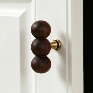 May include: A dark brown wooden door handle with three stacked spherical elements. The handle is mounted on a white door and features a brass-coloured base. The design is modern and minimalist.