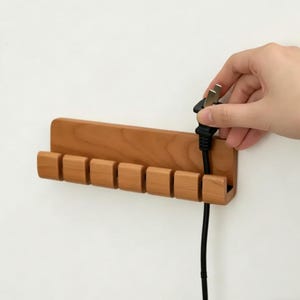 May include: A wooden wall-mounted organiser with seven slots, designed to hold plugs. The organiser is made of light brown wood and is mounted on a white wall. A black plug is being inserted into the organiser.