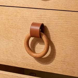 May include: A close-up of a wooden drawer pull with a circular wooden handle and a brown leather strap. The drawer pull is attached to a light-colored wooden drawer. The design is simple and modern.