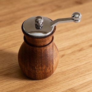 May include: A wooden coffee grinder with a stainless steel top and handle. The grinder has a dark brown, pear-shaped body and sits on a light brown wooden surface. The handle is attached to the top with a small knob.