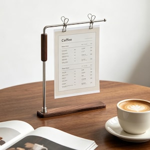 Könnte beinhalten: Ein Display-Ständer aus Metall und dunklem Holz mit einer horizontalen Stange und zwei silbernen Clips, die eine rechteckige Papierkarte mit der Aufschrift Coffee halten. Der Ständer hat einen rechteckigen Holzfuß und einen vertikalen Stab.