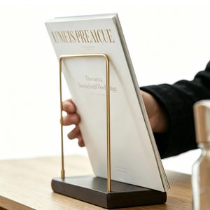 Puede incluir: Un moderno revistero con base de madera marrón oscuro y marco metálico dorado. Sostiene una revista con el texto "UNEIS PREAICUE" en letras doradas. El diseño es minimalista y elegante.