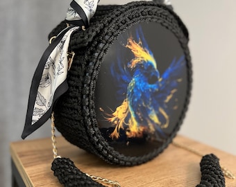 Pode incluir: Uma bolsa redonda de crochê preta com uma alça de tecido estampada em preto e branco e uma alça de corrente dourada. A frente da bolsa apresenta um design abstrato colorido de um pássaro.