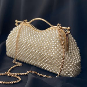 Pode incluir: Uma bolsa de m&atilde;o branca de p&eacute;rolas com uma al&ccedil;a de corrente dourada e um fecho dourado.