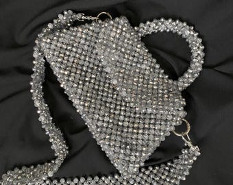 Pode incluir: Uma bolsa de mão de contas prateadas com alça superior e uma alça longa para o ombro. A bolsa é feita de pequenas contas redondas que são tecidas juntas para criar um padrão de malha.
