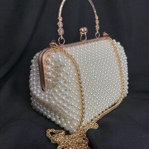Pode incluir: Uma bolsa de m&atilde;o de contas de p&eacute;rolas brancas com uma al&ccedil;a de corrente dourada e uma al&ccedil;a superior dourada.