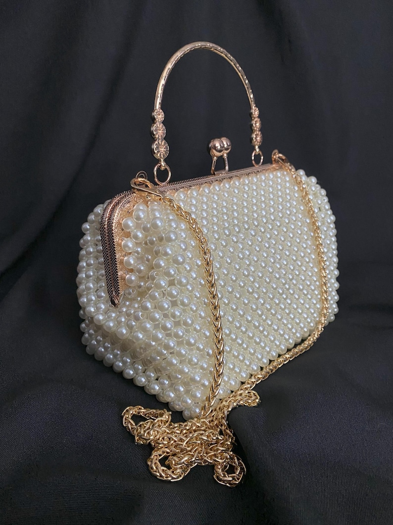 Pode incluir: Uma bolsa de m&atilde;o de contas de p&eacute;rolas brancas com uma al&ccedil;a de corrente dourada e uma al&ccedil;a superior dourada.