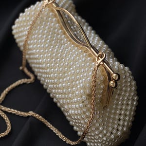 Pode incluir: Uma bolsa de m&atilde;o branca de p&eacute;rolas com uma al&ccedil;a de corrente dourada e um fecho dourado.