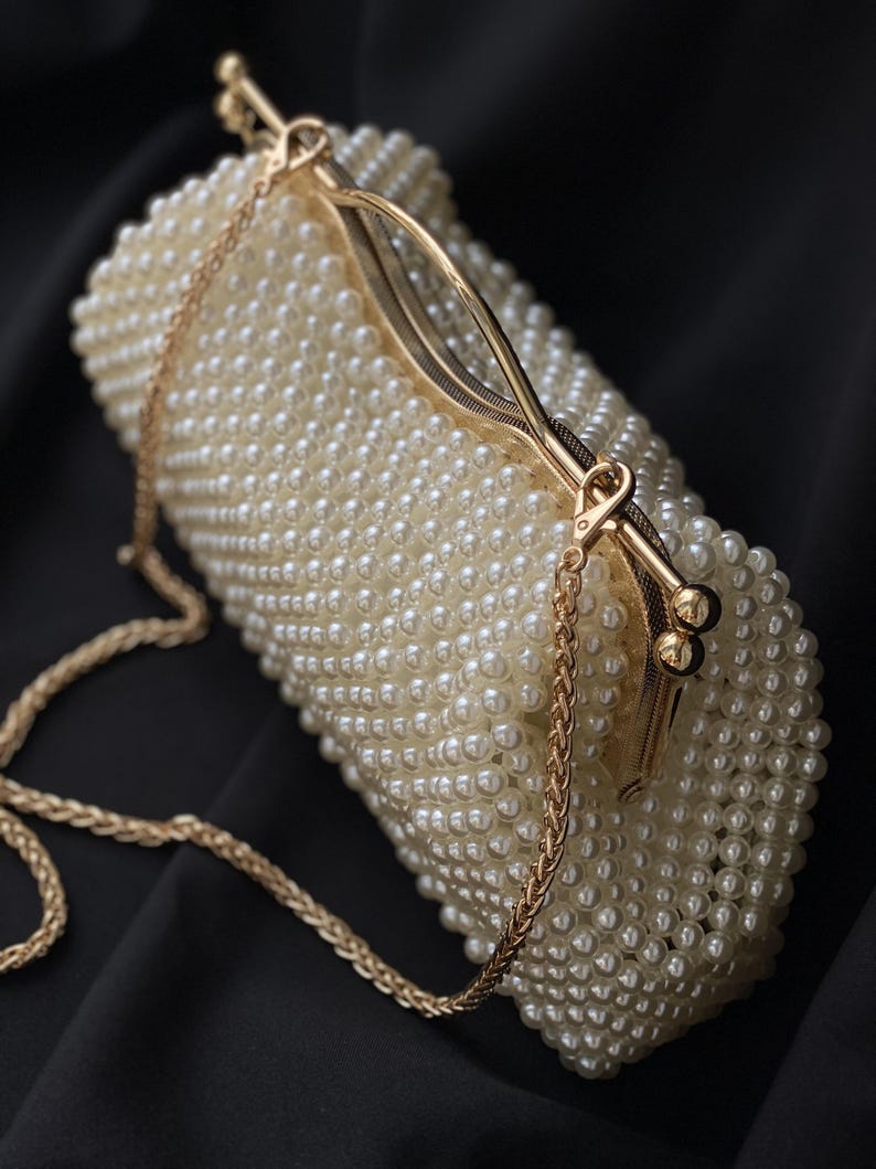 Pode incluir: Uma bolsa de m&atilde;o branca de p&eacute;rolas com uma al&ccedil;a de corrente dourada e um fecho dourado.
