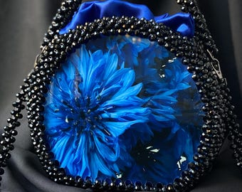 Pode incluir: Uma bolsa única, artesanal, com uma vibrante flor azul envolta em um corpo transparente e arredondado. A bolsa é adornada com contas pretas e tem uma alça e uma alça de contas combinando. O interior é forrado com tecido azul.