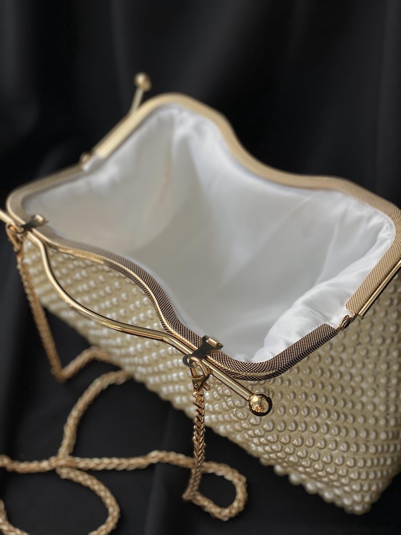 Pode incluir: Uma clutch branca de contas de p&eacute;rola com uma al&ccedil;a de corrente dourada e um fecho dourado. A clutch est&aacute; aberta e o interior &eacute; forrado com tecido branco.