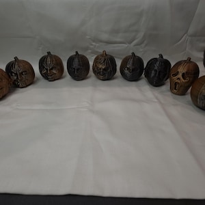 May include: A collection of ten small, round, pumpkin-shaped ornaments arranged in a horizontal arc. Each pumpkin features a unique, detailed sculpted face in either a metallic bronze or matte black finish, showcasing various horror-themed expressions.