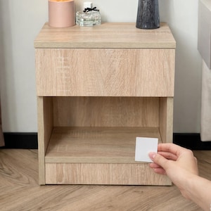 Pode incluir: Uma mesa de cabeceira de madeira clara com uma gaveta e uma prateleira aberta. Uma pequena lâmpada rosa com uma lâmpada redonda fica em cima, junto com um frasco de perfume e um vaso de lavanda. Uma mão segura um pequeno cartão branco perto da mesa de cabeceira.