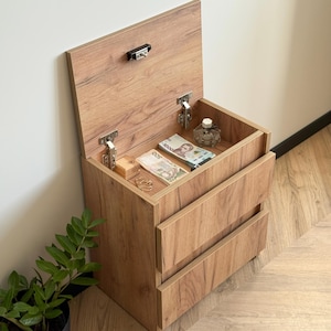 Pode incluir: Uma mesa de cabeceira de madeira com um compartimento superior articulado e duas gavetas. A parte superior está aberta, revelando dinheiro, joias e um frasco de perfume. A mesa de cabeceira é de madeira castanha clara.