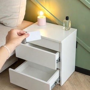 Pode incluir: Uma mesa de cabeceira branca com duas gavetas, uma aberta. Uma pequena lâmpada rosa com uma lâmpada acesa e um frasco de perfume transparente estão em cima. Uma mão segura um cartão branco perto da gaveta aberta.