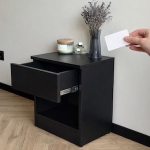 Pode incluir: Uma mesa de cabeceira preta com uma gaveta aberta, uma prateleira inferior e uma superfície superior. Um vaso com lavanda, uma vela e um frasco de perfume estão no topo. Uma mão segura um cartão branco perto da mesa de cabeceira.