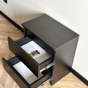 Pu&ograve; includere: Un comodino grigio scuro con due cassetti aperti, che rivelano interni bianchi. Il cassetto superiore contiene piccole piastrelle bianche e un pacchetto con un'etichetta rossa e verde. Il comodino &egrave; su un pavimento in legno chiaro.