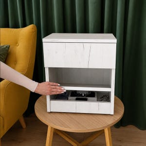 May include: A white wooden bedside table with a wood grain texture, featuring a top drawer, an open middle shelf, and a bottom storage compartment. The unit has a rectangular shape and is constructed from light-coloured wood panels with visible grain.