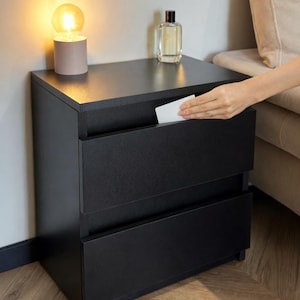 May include: A black nightstand with two drawers. A small lamp with a round bulb and a perfume bottle sit on top. A hand is reaching into the top drawer. The nightstand is placed next to a beige sofa.