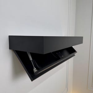 以下が含まれることがあります： 黒色の壁掛けキャビネット。蝶番付きのドアが部分的に開いています。長方形のキャビネットは白い壁に取り付けられています。ドアは金属製の腕で支えられています。