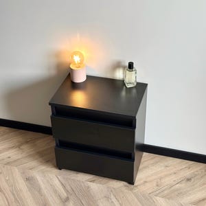 Pu&ograve; includere: Un comodino nero con due cassetti. Una piccola lampada rosa con una lampadina accesa e una bottiglia di profumo trasparente sono sopra. Il tavolo &egrave; contro un muro bianco e su un pavimento in legno.