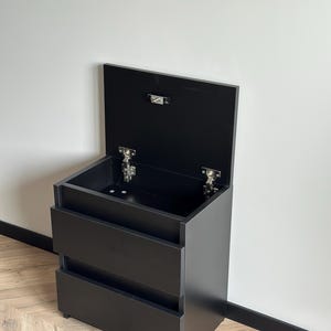 Pu&ograve; includere: Un mobile contenitore nero con due cassetti e un coperchio incernierato. Il coperchio &egrave; aperto, rivelando l'interno. Il mobile &egrave; realizzato in un materiale scuro e opaco, con cerniere e un fermo argentati.