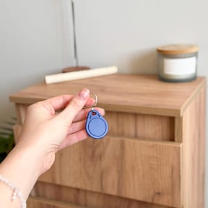 Pode incluir: Um porta-chaves azul em forma oval com um porta-chaves prateado. O porta-chaves é segurado em frente a uma cômoda de madeira. Uma vela em um frasco de vidro com tampa de madeira está na cômoda.