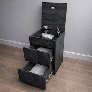 Pode incluir: Um armário cinza escuro, com textura de pedra, com duas gavetas e um compartimento superior. A parte superior está aberta, revelando um pequeno objeto no interior. As gavetas estão parcialmente abertas, mostrando interiores brancos. O armário está sobre um chão de madeira.